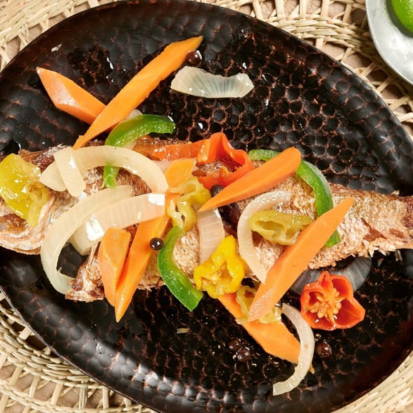 Whole fried fish topped with colorful bell peppers, onions, and carrots on a dark plate.